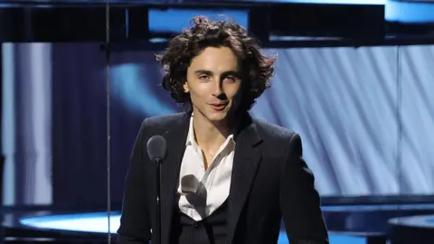 Picturegroup Timothée Chalamet wears a navy three-piece suit with a white shirt open at the neck. He's standing in front of a light blue patterned stage backdrop and speaking into a microphone on a thin stand. He looks relaxed and is smiling as his medium-length black hair cascades down, framing his face.