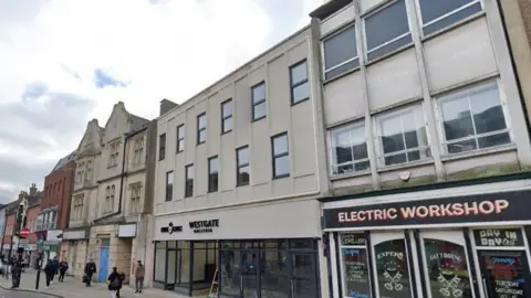 Westgate, Peterborough: The side of a street. Shoppers are walking past buildings in a street. Some building has names and signs written on them. One the right is a sign saying "ELECTRIC WORKSHOP". In the centre is a sign saying "WESTGATE"