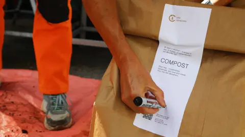 Heledd Wyn A large paper sack with a white label on the outside reading Compost. There is a Criw Compostio logo at the top and smaller typing plus a bar code. One of ffin's legs wearing orange trousers is visible behind it, and a hand holding a permanent marker bending over to write something on the label.