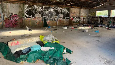 Interior of a large abandoned warehouse with graffiti-covered walls and exposed metal roof beams. The concrete floor is scattered with rubbish, clothing, and discarded materials. In the foreground, torn green tarpaulins and fabric lie beside a low concrete structure with graffiti. Large black-and-white mural artwork covers part of the back wall, while daylight enters through windows on the right side of the building.