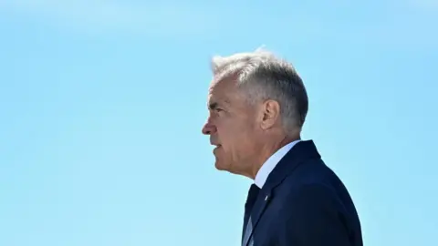 Mark Carney stands in profile, facing to the left, against a clear light blue sky. He wears a dark blue suit and tie.
