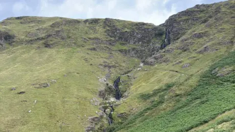 A narrow waterfall carved into the side of a steep and rocky fell. The slope is covered in grass with various rock outcrops. The waterfall curves down the side of it. The sky is blue with thin clouds.
