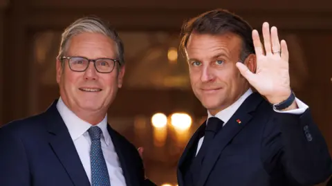 UK Prime minster Sir Keir Starmer and French President Emmanuel Macron stand in the doorway of number 10 Downing Street. President Macron is waving.