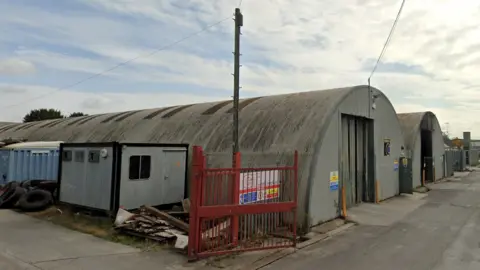 Two buildings on an industral estate. The buildings are tube shaped and grey. There is a red fence in the foreground and an electricity pylon.