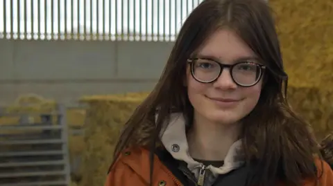 Molly-Mae stands in a barn in front of a stack of hay bales. She has long brown hair and glasses and is smiling. She is wearing an orange mac