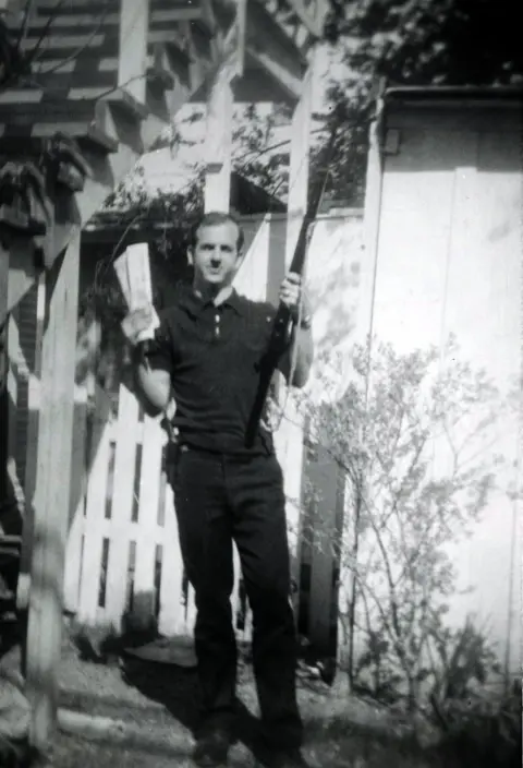 Reuters/Dallas Police Department A black and white and slightly blurry image of Oswald holding a gun and papers, taken from a distance, with a fence and stairs in the background