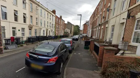 A Google street view of Cromwell Street in Gloucester with cars parked on one side, and a row of terraced houses.