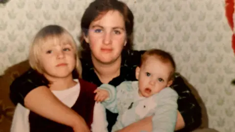 Lilly family A photograph taken in the 1980s, showing Vicky as a baby in her mothers arms, and she has her right arm around Sarah, who has short blonde hair