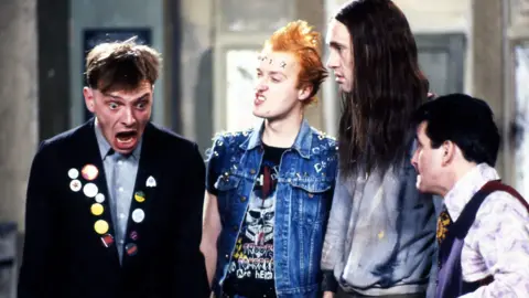 BBC An image from sitcom The Young Ones, showing Mayall, Ade Edmondson, Planer and Christopher Ryan. Mayall, in character wearing a dark jacket including badges, is looking shocked and the others are looking at him.
