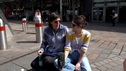 Two people sit on a seat along a pedestrian street. One is wearing a blue tie dye hooded top and sunglasses, and the other is wearing a white, yellow and lilac stripe cardigan and sunglasses..