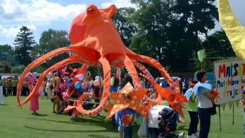 Image of a large orange fabric octopus puppet being held in the air by poles by a group of festival goers. In the foreground is a woman holding a banner in the background can be seen crowds of people, trees and balloons.