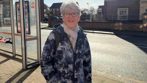 A woman with white short hair and sun in her eyes, smiling at the camera in front of a bus stop