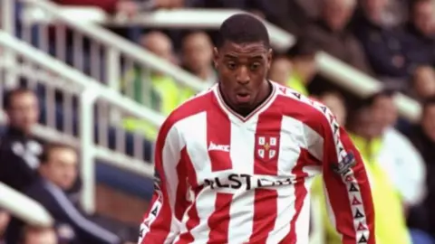 AllsportUK/Getty Images A man with close-cropped black hair during a football match in front of a stand full of spectators. He is wearing a football shirt with vertical red and white stripes and the badge of Lincoln City FC.