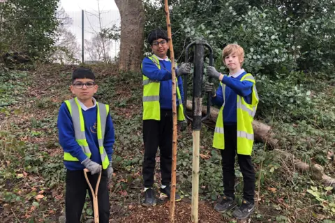 BBC / Naj Modak Three children in blue pullovers and black trousers, the boy on the right is holding a spade while the other two stand beside a newly planted tree