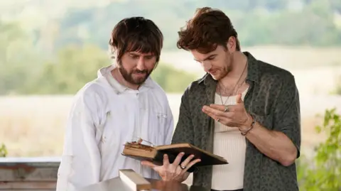 James Buckley and Jonno Davies stand outdoors looking at an open book.