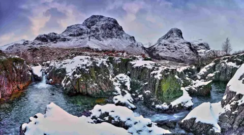 John Cuthbertson Water flowing between rocky ground covered in snow with the Three Sisters of Glencoe in the distance