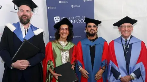 University of Liverpool handout Left to right: David Winstanley, Wendy Beetlestone, Ringo Starr and alumnus Brian Boyd. They are all wearing graduate robes with mortarboard caps. They are standing in front of a University of Liverpool exhibition board. They are all smiling.
