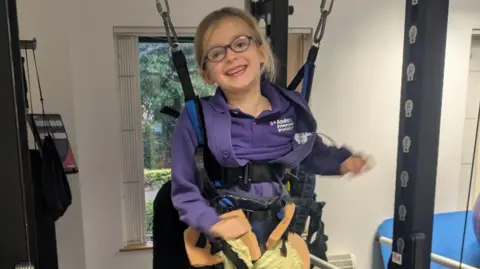 PA Media Sofia Hill during a physical rehabilitation session. She is pictured in a hoist and is smiling at the camera. 