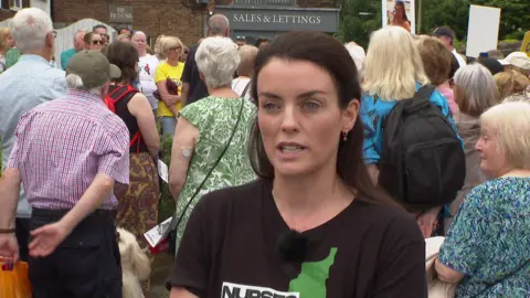 Lucy Williams, who has straight brown hair and wears a black t-shirt, speaks to the camera, while a large group of campaigners gather in the backdrop