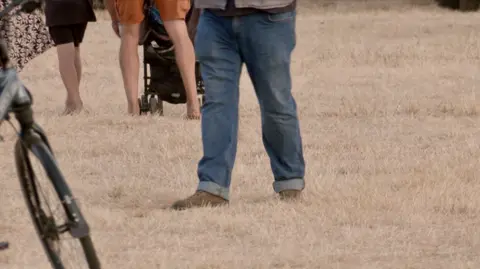 A close-up of dry, yellow grass on a field. Several people's legs are visible as they stand on the grass.