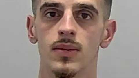 Nardi Bytyci: A man with short dark hair, a dark moustache and dark goatee beard looking ahead and wearing a grey round-necked top