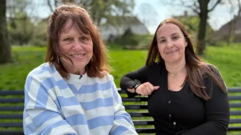 Margaret and Leanne sit on a bench in a park. Both are smiling at the camera. Margaret has short dark hair, and wears a white and purple striped jumper with a gold locket necklace. Leanne has longer dark hair, and is wearing a black cardigan, a black watch and a short silver necklace. 