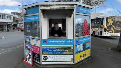 States of Guernsey A woman leans over the open hatch of a hexagonal kiosk at a bus terminal. It has the logo of the Royal British Legion on it and a bus is driving by to the right of the kiosk.