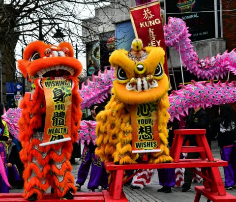 Raymond Watson Orange and yellow Chinese dragons with pink tails stand on red structures during a street performance. The dancers' purple trousers can be seen below the huge puppets. 
