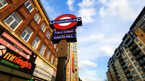 A street-level view of the London Underground sign for Gants Hill station, featuring the red and blue roundel against a bright blue sky with scattered clouds. Surrounding the sign are multi‑storey brick and modern buildings, including shops with colourful signage along the street.