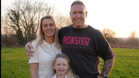 A mother, father and young girl with blonde hair posing outside.