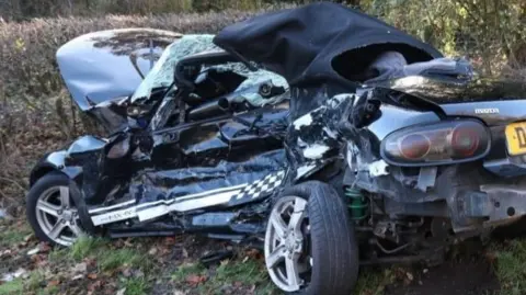 The Air Ambulance Service A very badly damaged car, with a smashed windscreen, the passenger side bent inwards and the rear tyre off the wheel. The car is pointing into a hedge