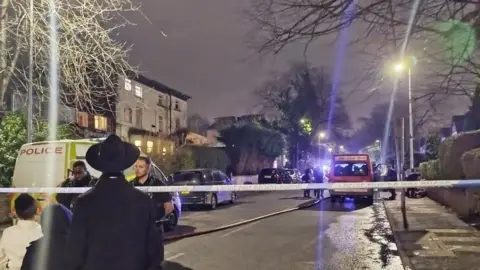 Police tape cordons off scene of fire. Two boys and a man stand on one side with a police van the other side and a red van in the distance 