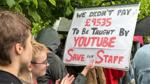 Students standing in the rain at a protest. One is holding up a sign saying: 'We didn't pay £9,535 to be taught by YouTube. Save our staff."