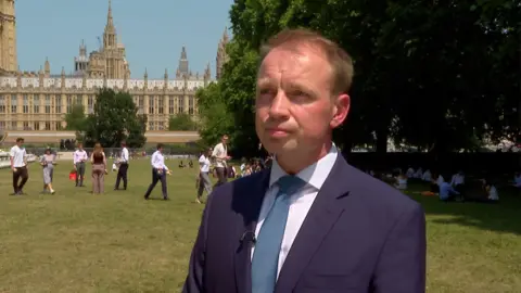 A man is stood in a park in London. The Houses of Parliament are in the background. he is wearing a navy blue suit and a blue tie. he has brown hair.