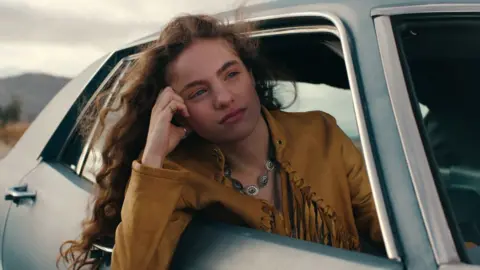 Netflix Autumn Best dans le rôle d'Amy dans Woman of the Hour. Elle est penchée du côté passager de la voiture et porte une veste en daim couleur moutarde avec des pompons. 