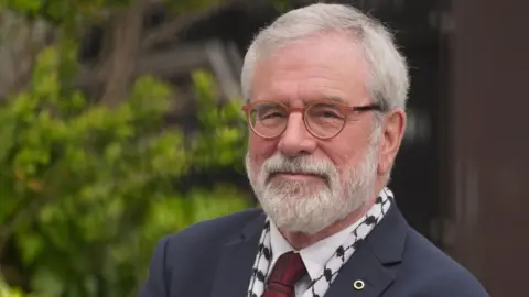Gerry Adams -  a man with grey hair and beard - is smiling at the camera, wearing a navy suit jacket white shirt and red tie. 