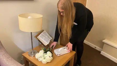 Becca Cook, a young woman with long blonde hair and black glasses, reorganises a table in a chapel of rest. On the table is a bunch of fake white roses, a box of tissues, a lamp with a beige shade and a religious book which is open on a stand. She is wearing a black dress with black tights. The carpet is brown.
