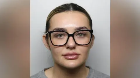 A mugshot of a woman looking at the camera. She is wearing thick-rimmed dark glasses and has her hair tied back. 