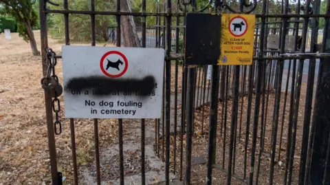 Gloucester City Council A photo of two warning signs in Gloucester Old Cemetery. One reads no dog fouling in cemetery and the second tells dog owners to clean up the pet's mess. The signs are fixed to some black railings. The signs have black spray paint covering the message to keep dogs on leads.