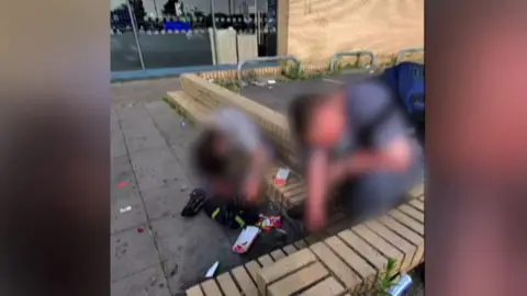 Two people sit on a wall, surrounded by items on the ground. The image has been blurred to protect the identity of the individuals. 
