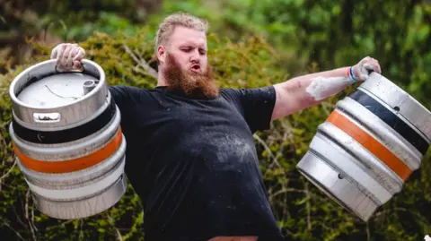 Barley Mow A man in a black t-shirt with a large beard and light hair is holding a barrel of beer in each hand. He is outside standing by a hedge.