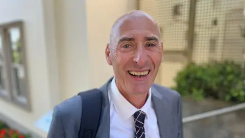 Deputy Steve Williams smiles into the camera. He is wearing a grey blazer and a white shirt with a navy tie.