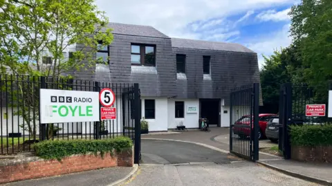 BBC Radio Foyle's building in Londonderry has a large black gate outside it. There is two Radio Foyle signs on either side of the gate.
A number of trees are also visible on the grounds. 
