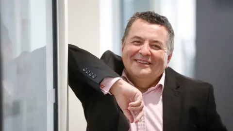 Andy Barnes Richard Skellett, wearing a dark jacket and pink shirt, smiles while looking into the camera