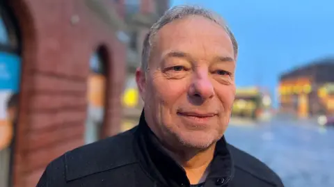 Blackpool and Fylde Street Angels founder Paul Rawson standing outside the Clifton Street building in the early morning. He has short grey hair, wearing a dark coat with the collar up and is smiling. Blackpool town centre can be seen blurred in the background 