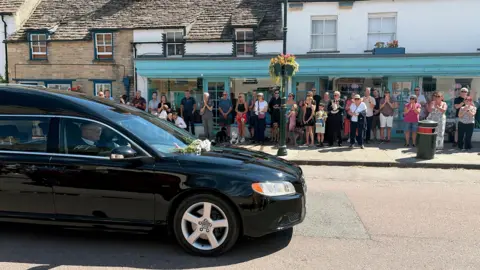 PA Media A black hearse in a historic street on a sunny day. People line up along the pavement.