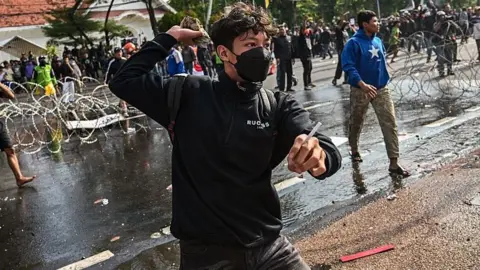 AFP via Getty Images A young man, wearing a black face mask and a rucksack, appears to hold a rock back ready to throw on a busy road during demonstrations