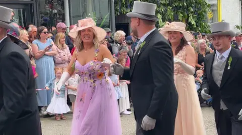 Four couples are dancing in the street, wearing mornign coats and smart frocks . The pavements are lined with spectators.
