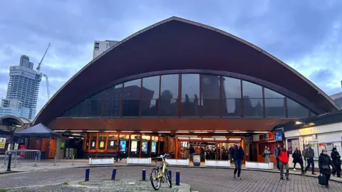 The timber facade of Manchester Oxford Road looms above the entrance to the station. Passengers can be seen walking out from the ticket stiles below on to a paved concourse. Several high-rise buildings can be seen in the distance on a cloudy day.