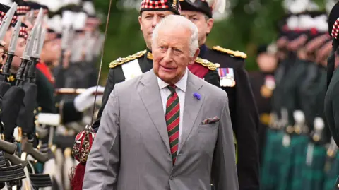 King Charles III wearing a grey suit with a white shirt and red and green striped tie. He is walking through the Palace Guard - a line of soldiers, wearing black uniforms.
